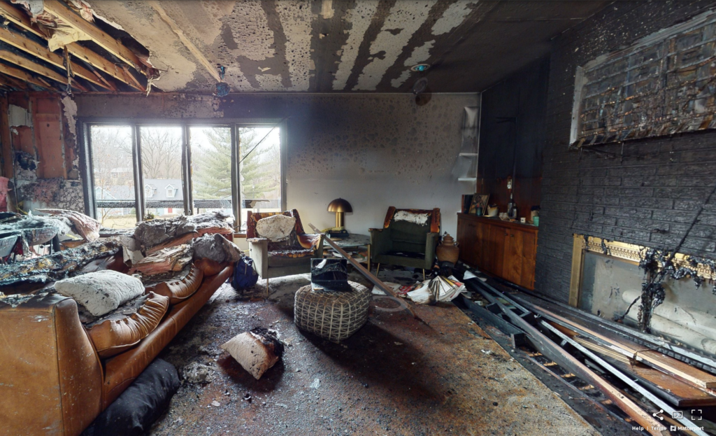 Burned living room with charred furniture, smoke-stained walls and ceiling, and debris scattered across the floor