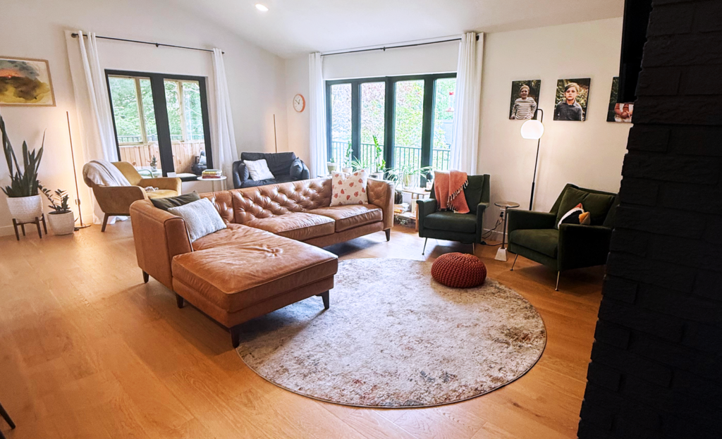 A bright living room with a tan sectional sofa, green chairs, large windows, and framed portraits on the wall